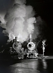 two steam locomotives waiting at a depot at night